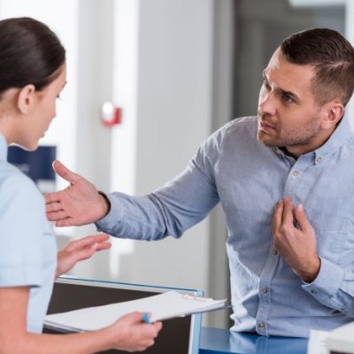 A man looking aggressive and threatening stands before a woman at a medical reception desk.